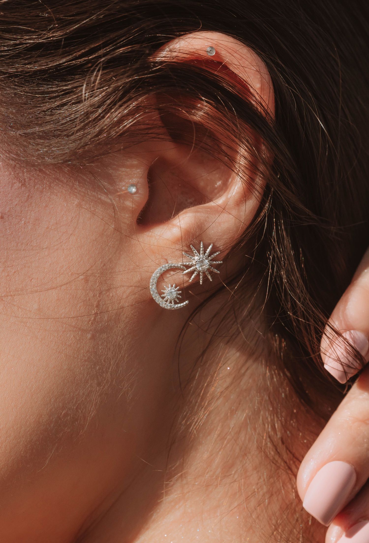 A sunlit ear with a coordinated celestial set: a pavé crescent-moon climber, a starburst stud, and a helix stud, an ear composed over time.