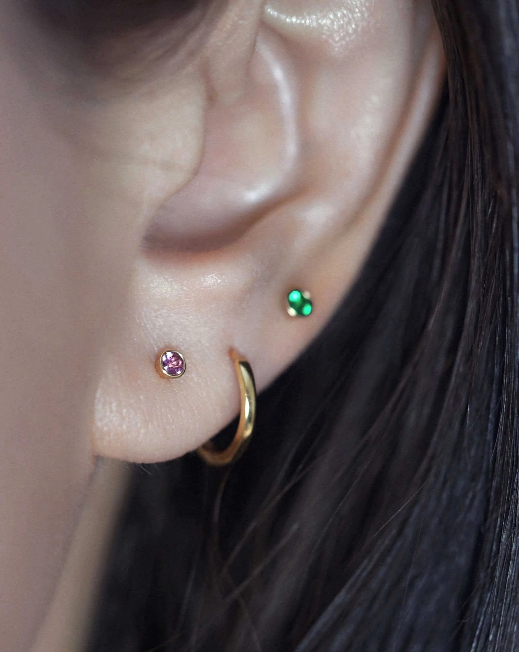 A lobe wearing a pink gemstone stud, a gold huggie, and a green trinity stud, a deliberately asymmetric arrangement that reads as personal choices.