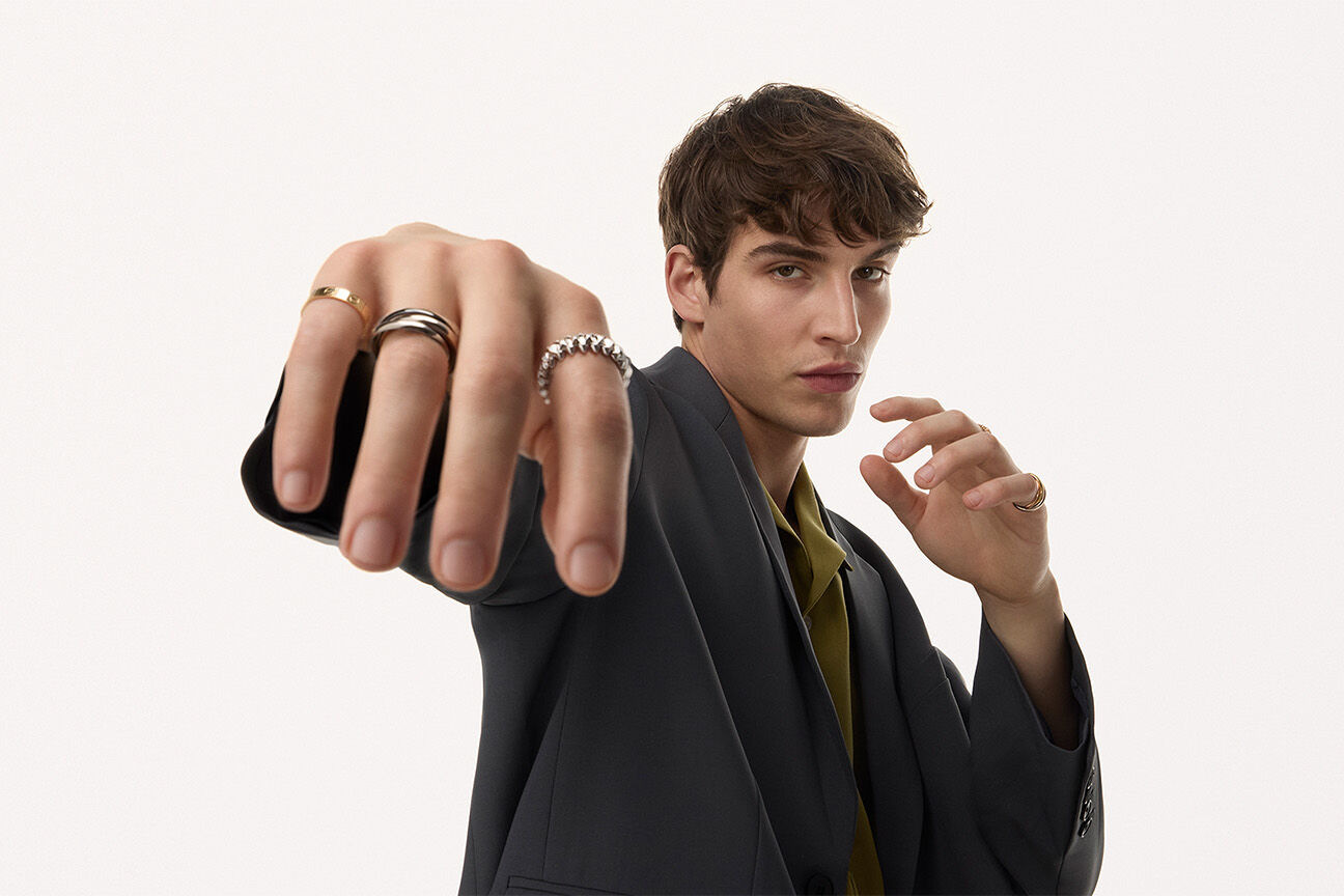The banner Cartier runs across its own rings-for-men page: a man in a charcoal suit, one hand thrust forward wearing a plain gold band, a three-band Trinity and a chain-link ring - every ring on that page sits on the main rings page too, at the same price.