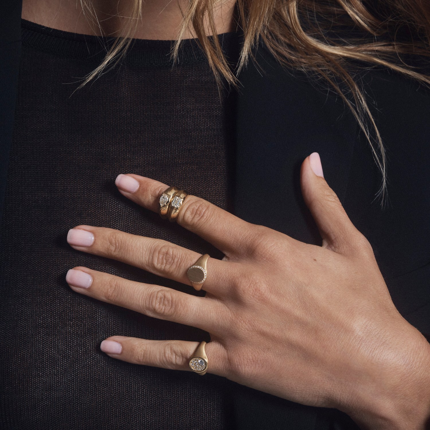 A woman's hand resting on a black knit sweater, wearing two Jade Trau Tatum signet rings, one pave-edged and one pave-set - a woman can wear a signet ring, the same fact as a man in a pearl necklace, running the other direction.