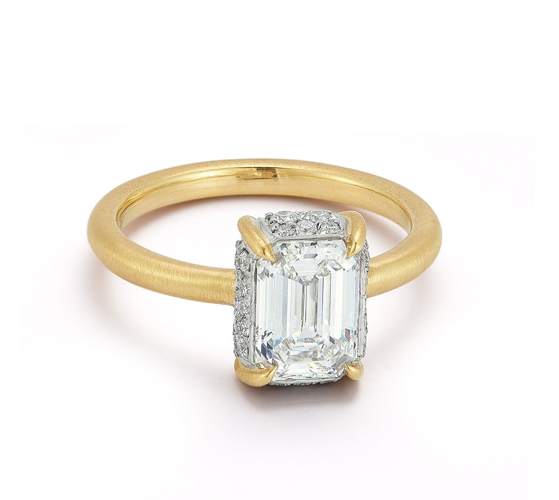 Jade Trau emerald-cut diamond framed by fine pavé and gold prongs on a brushed yellow-gold band: the signature setting that lets you read the stone's shape from across a room