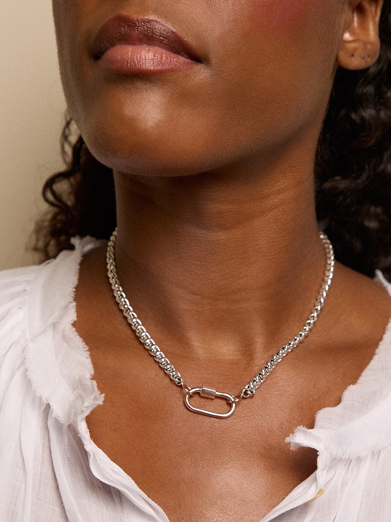 A two-tone Marla Aaron Lock, silver body with a gold closure, clipped onto a rolling-curb chain and worn at the collarbone, the carabiner-as-jewelry signature that sells through used and proof you don't need the all-gold version.