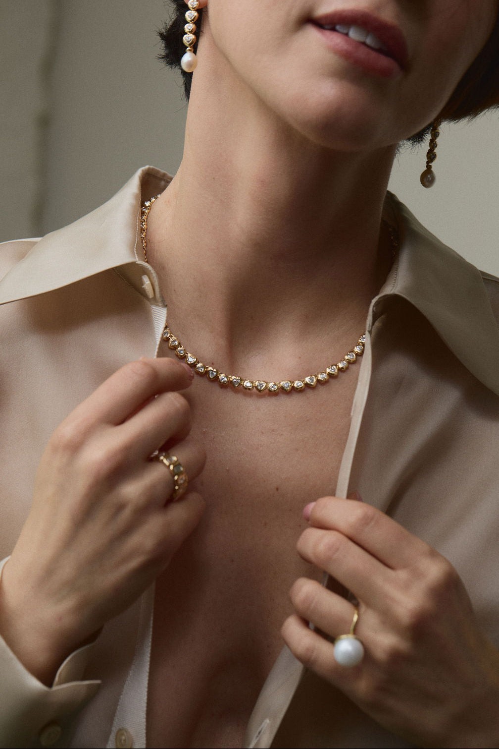 A delicate gold and white-sapphire riviere necklace and a few fine gold rings worn under an open cream silk shirt, the kind of signature fine jewelry whose retention on the resale market is read one piece at a time.