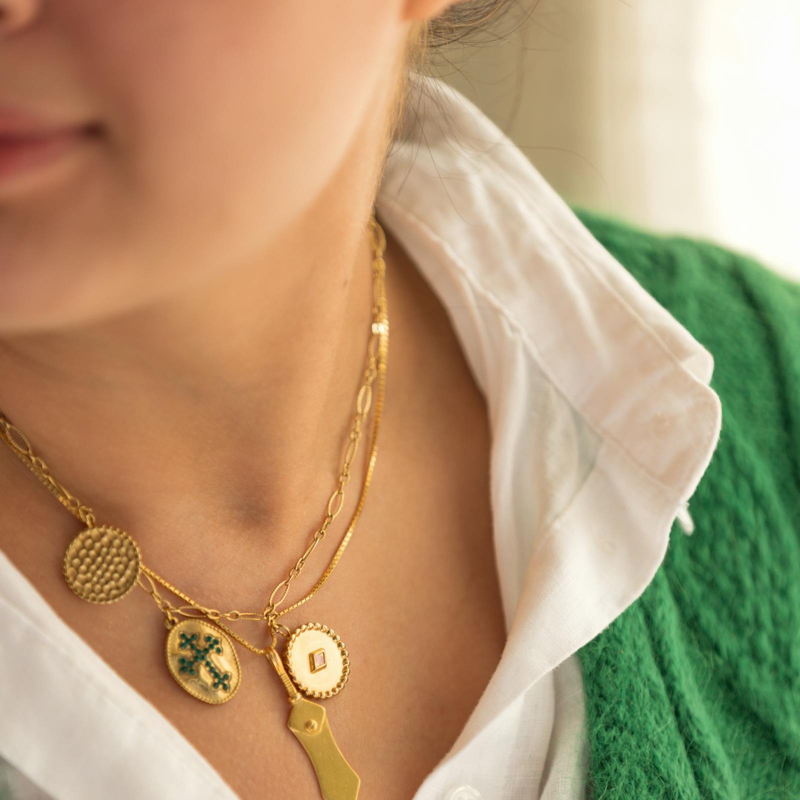 A layered medallion-and-charm neck stack on a white shirt and green knit, gold discs and engraved charms hung together, the kind of collected, expressive look the 2026 charm-piled-chain trend is built around.