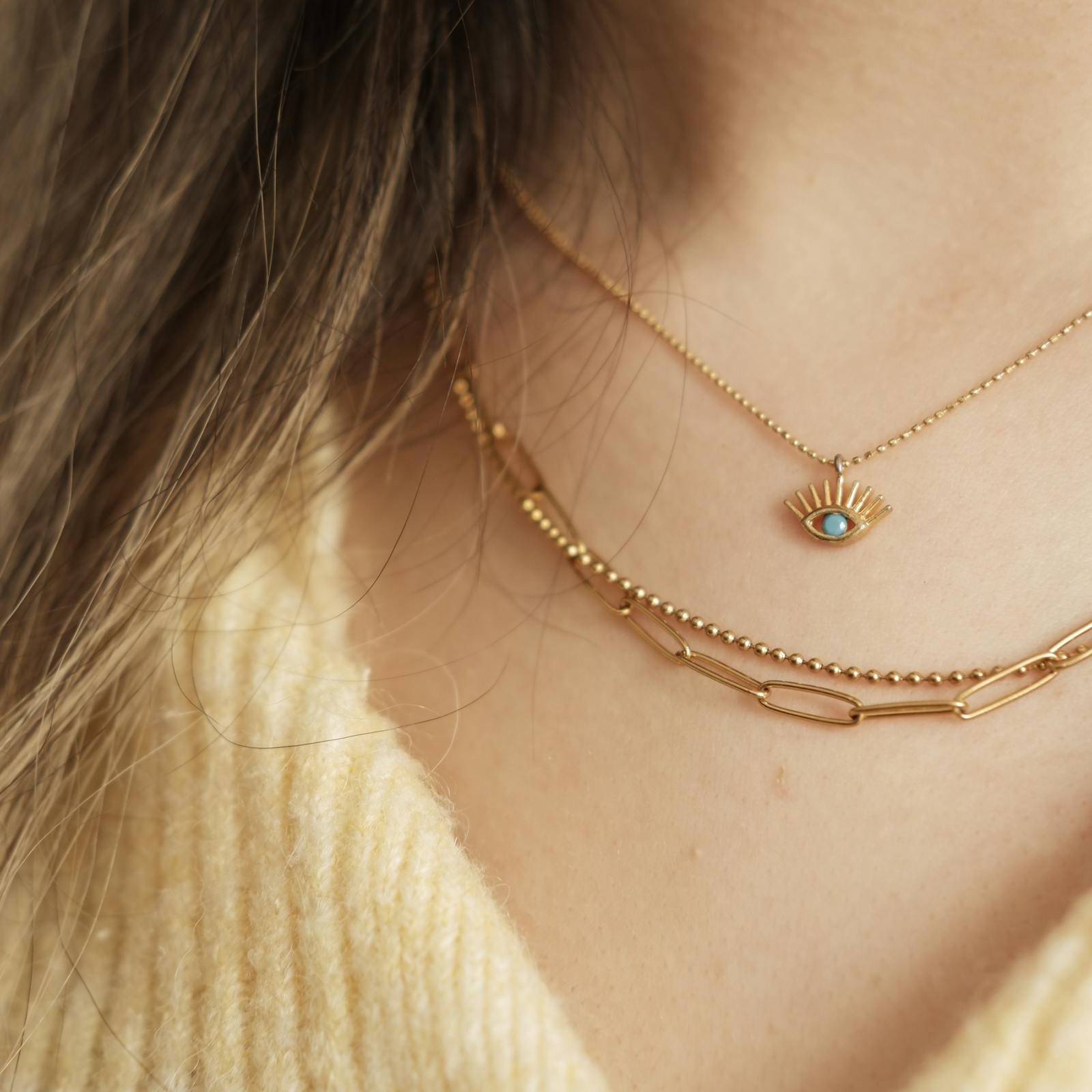 A three-piece gold neck stack worn on a butter-yellow knit: a fine ball chain with a turquoise evil-eye pendant, a beaded chain, and a paperclip chain. The kind of clean, deliberate stack a reader who knows can read across a room.