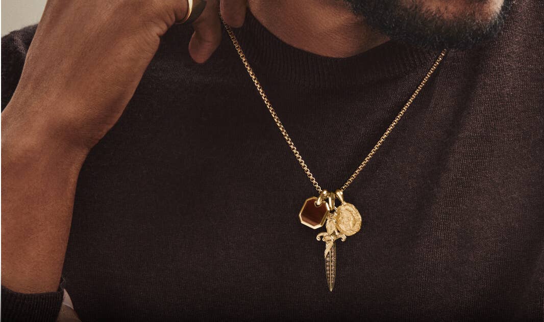 A man in a dark crewneck knit wearing a cluster of David Yurman gold amulets on a box chain.