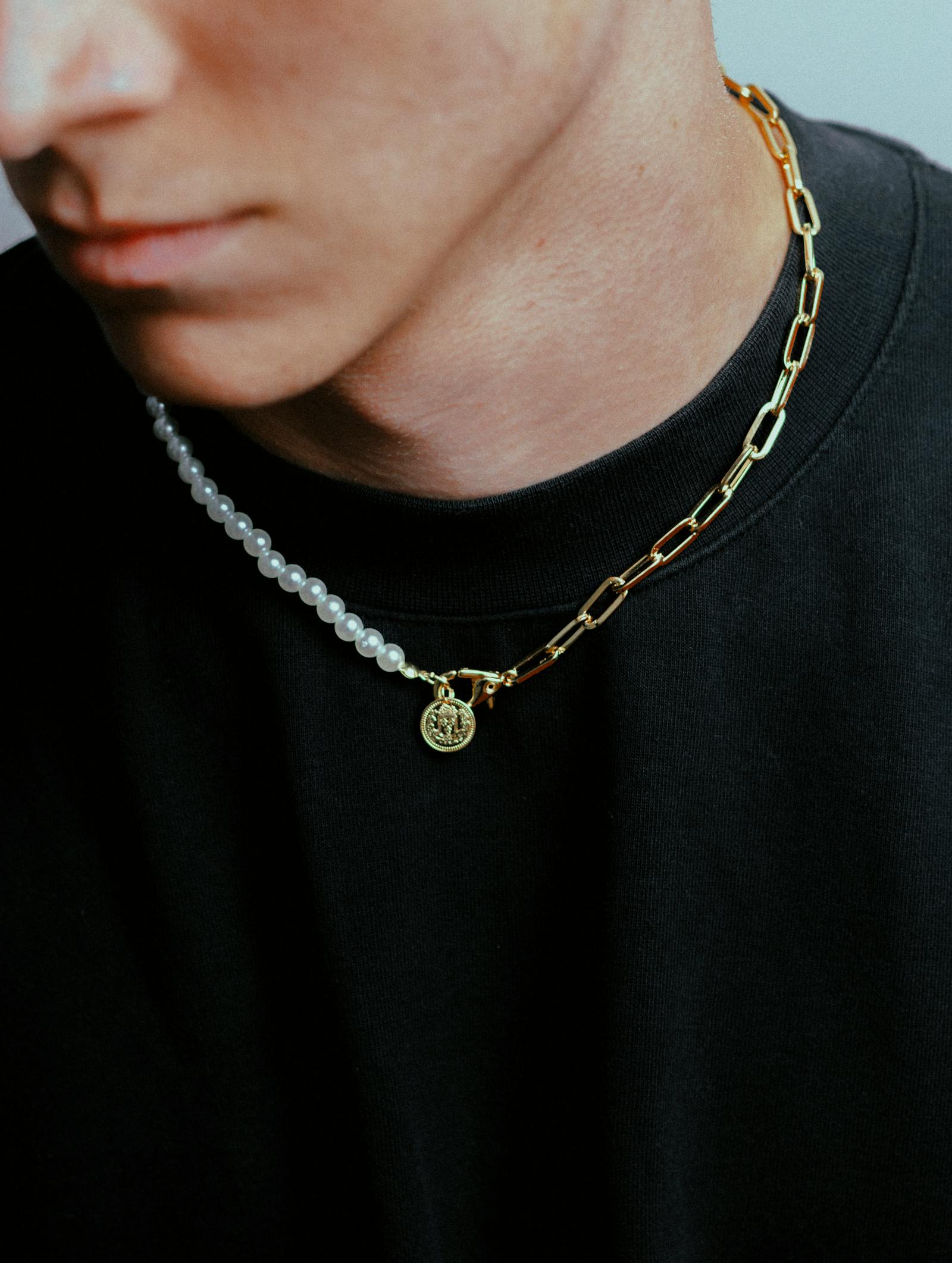 Close-up of a man in a black t-shirt wearing a small round gold coin medallion on a layered gold paperclip chain and pearl chain.