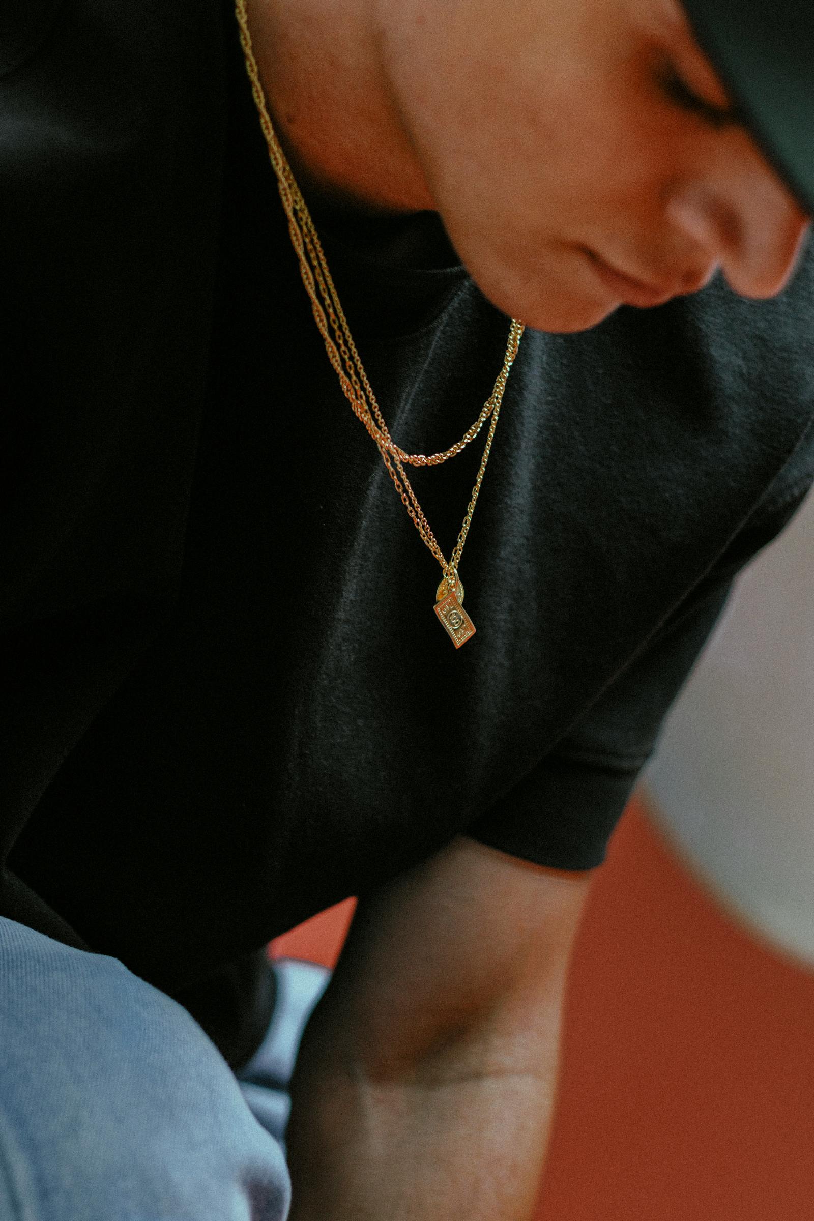 A man in a black t-shirt wearing layered fine gold chains with a small rectangular pendant.