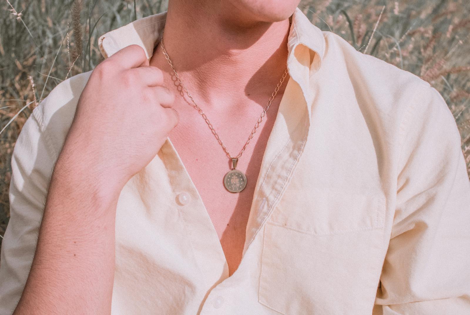 A man in an open cream linen shirt wearing a round vintage gold medal pendant on a fine chain, shot in a sunlit field.