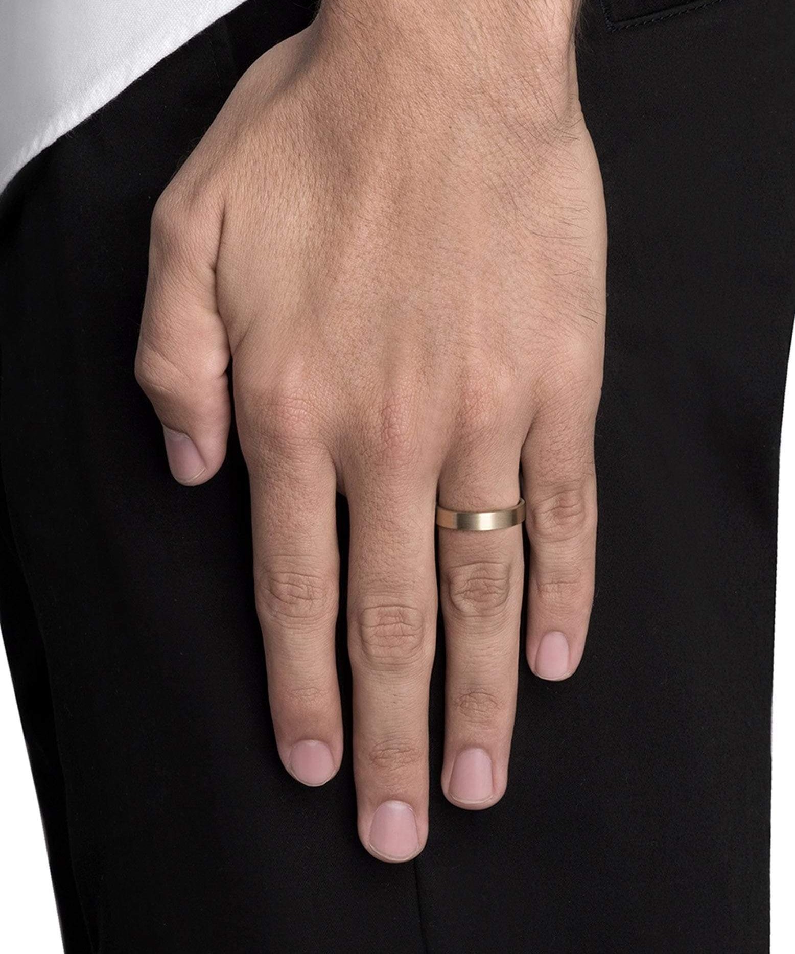 A plain matte gold band worn on a man's finger, not the ring finger, against black trousers, the solid gold band worn because you like it rather than because you're married