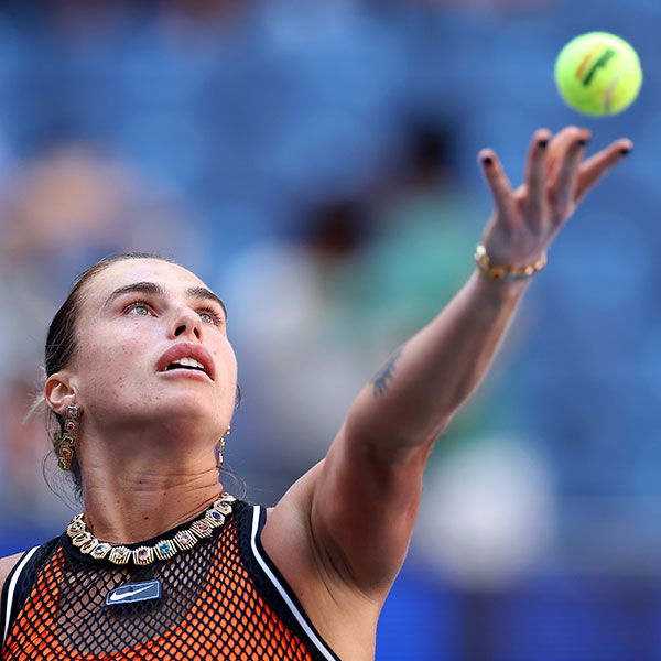 Aryna Sabalenka mid-serve at the 2026 US Open, eyes on the tossed ball, wearing Material Good's gold collar of single bezel-set colored stones and the matching bracelet on her serving wrist.