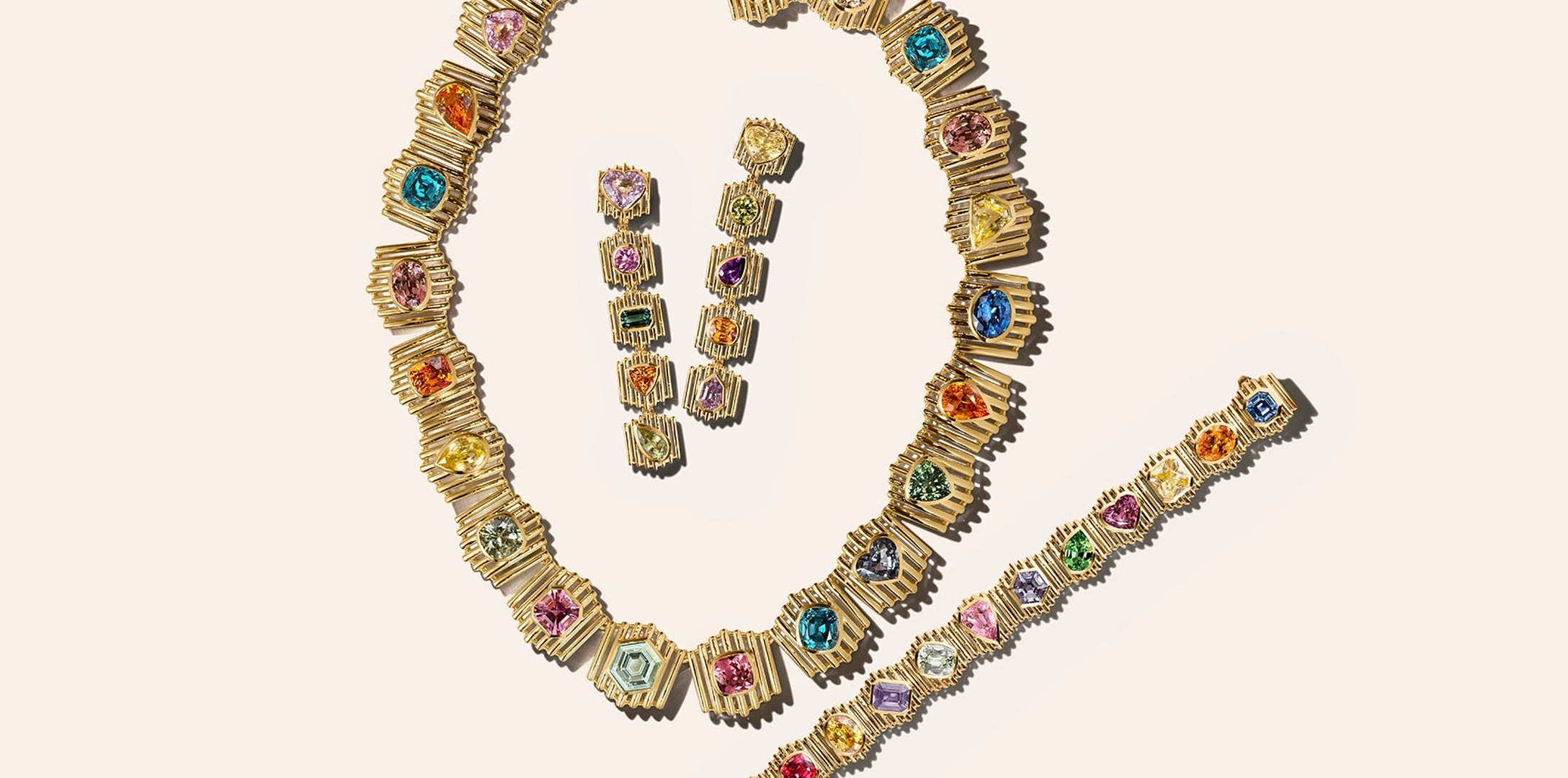 Material Good's Prism suite laid flat on cream: a necklace, a bracelet and two drop earrings of fluted gold links, each holding one colored stone in its own bezel with bare gold on either side. No pavé anywhere on the three pieces.