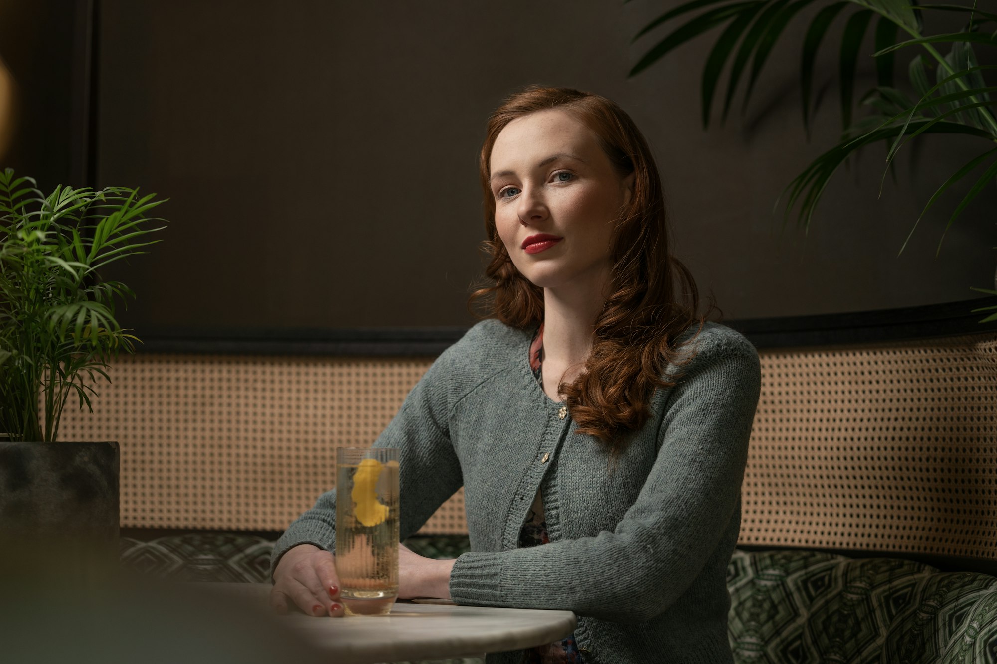 A woman sitting alone and composed at a bar, wearing fine jewelry, the picture of someone buying for herself without rehearsing a reason