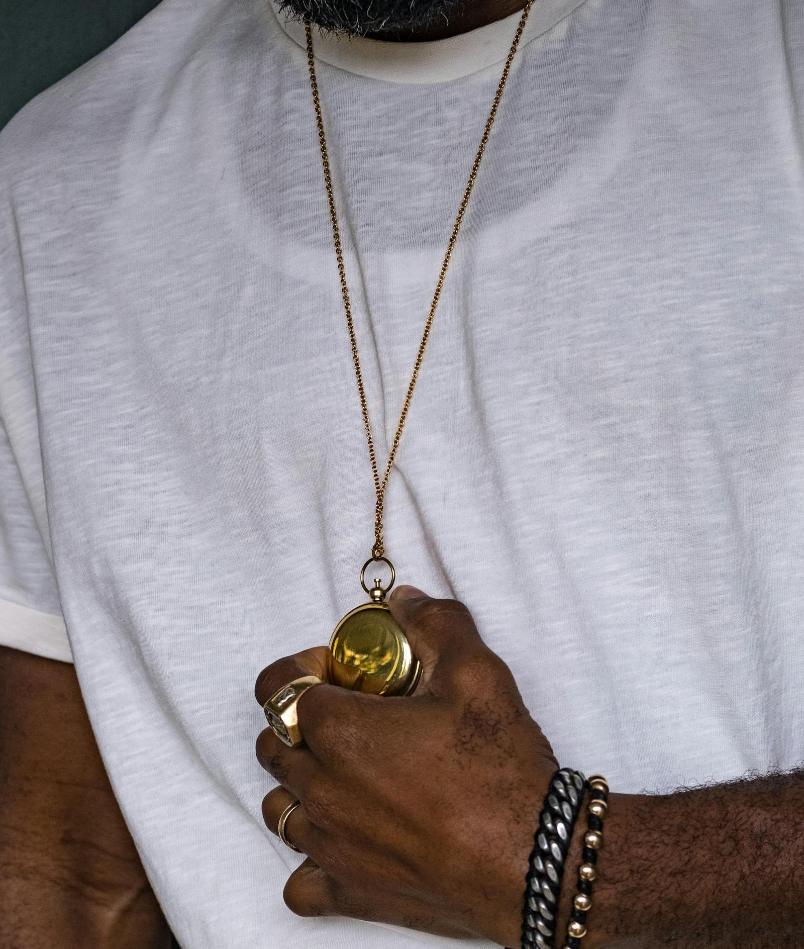 A man's hand holding his own gold medallion on a long chain, a signet ring and stacked bracelets on the same hand, the deliberate self-purchase of a male collector