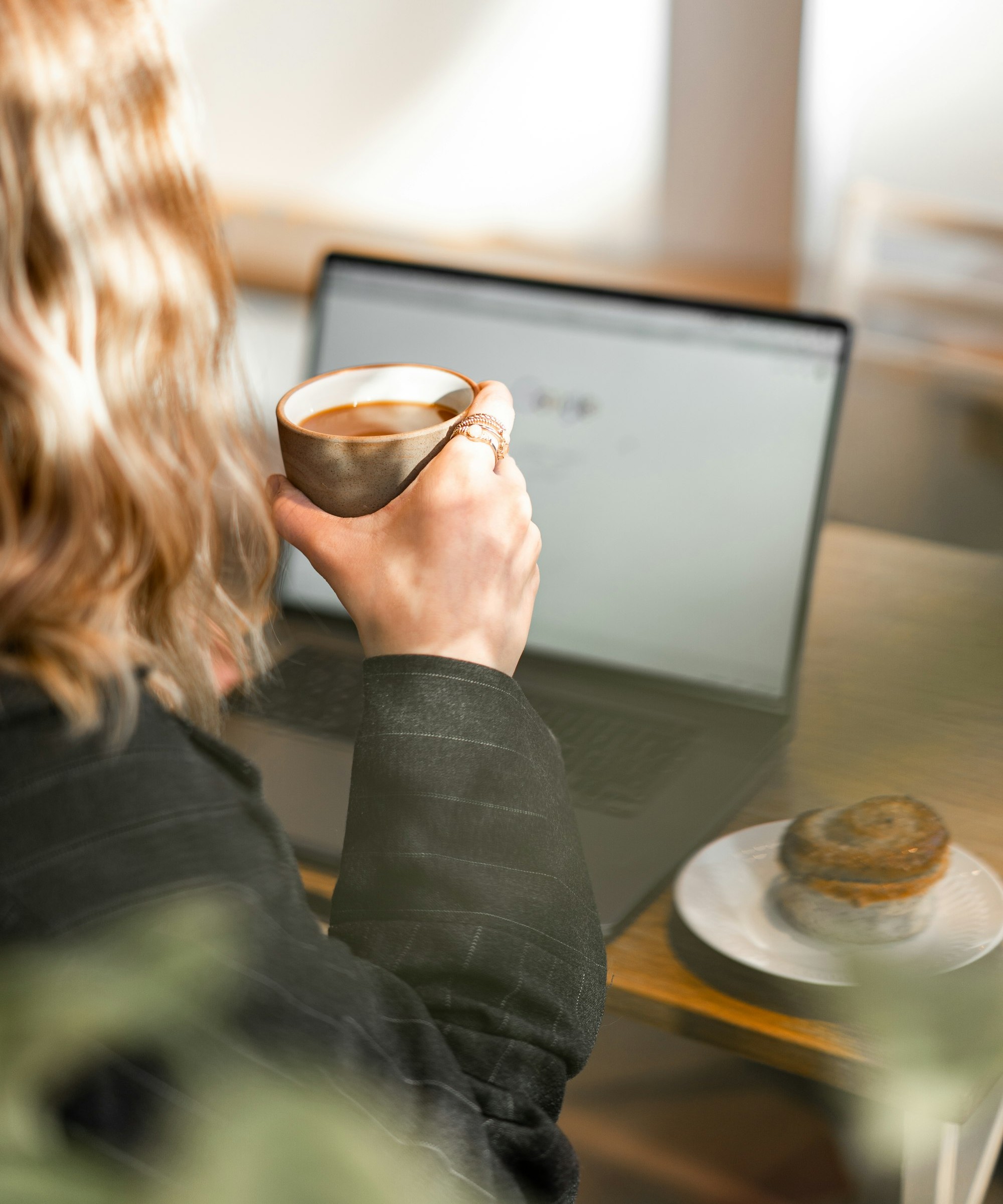 A woman with stacked rings on her own hand holding a coffee cup while a laptop sits open in front of her, the online research of someone buying fine jewelry for herself