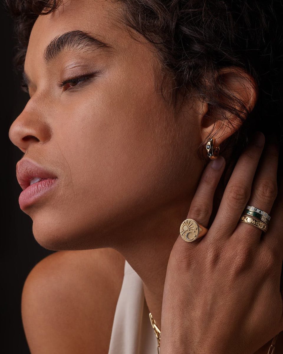 A woman wears a gold signet ring engraved with a sun and crescent moon on her finger, the same kind of gold signet a man might wear, which is why the form's 2026 moment belongs to women and men equally.