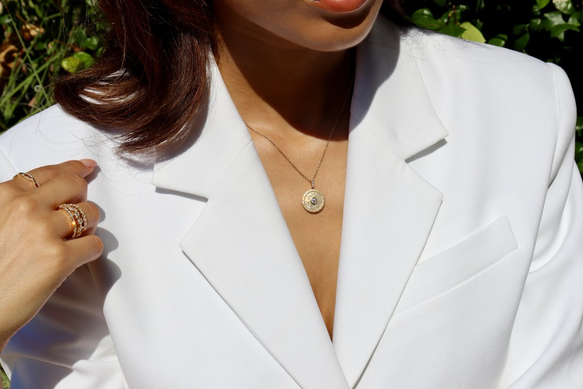 Gold disc pendant with scattered diamonds on a delicate chain, worn over a white blazer in natural light