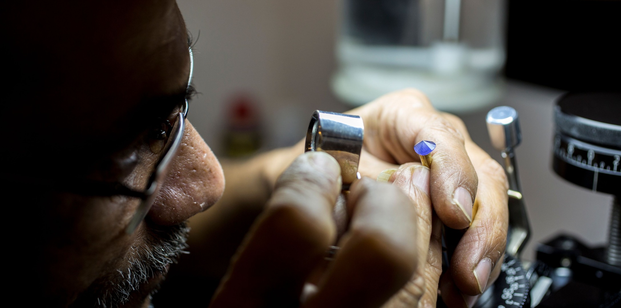 A man at a gem-testing bench reads a small blue faceted stone through a loupe, a refractometer at his elbow.