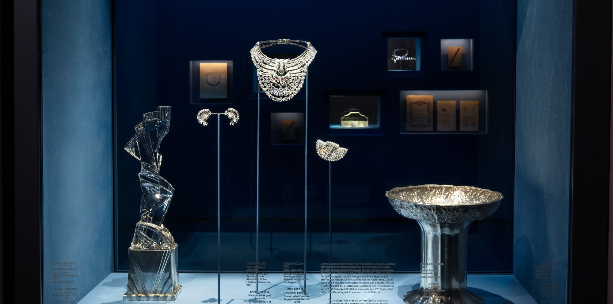 The 1939 Van Cleef & Arpels diamond necklace on its stand inside a lit blue vitrine at the MAK in Vienna, with earrings, a brooch and a silver bowl beside it. This is the case two men broke with a hammer on 27 August 2026.